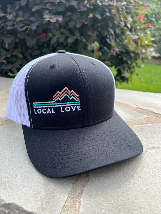 A black baseball cap with the words Local Love written in white.  Also includes a graphic with horizontal lines and lines that symbolize mountains. Back of cap is white mesh.