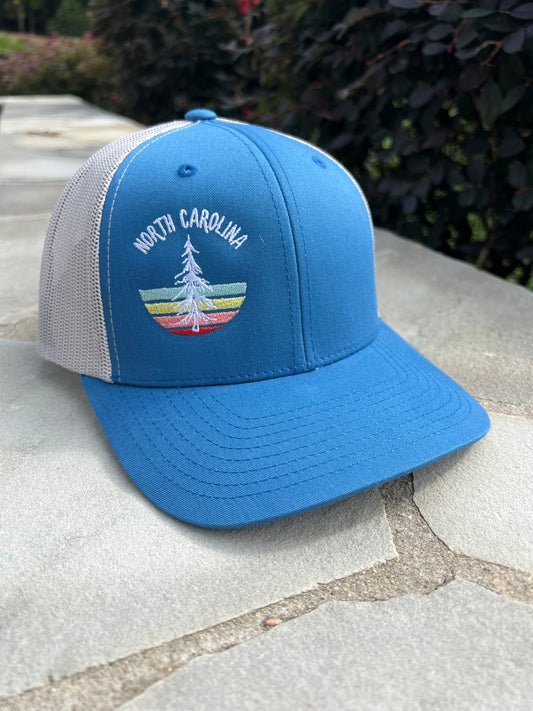 A blue baseball cap with North Carolina written on it in white lettering and a graphic of a white pine tree with red, pink, yellow and green stripes behind it. The cap also has a white mesh backing.