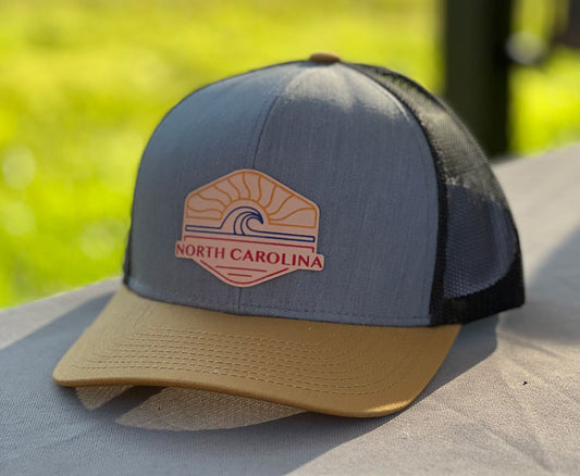 A baseball cap style hat with a tan brim, blue front, and black mesh back, with a  patch that has a sun and wave and North Carolina on it.